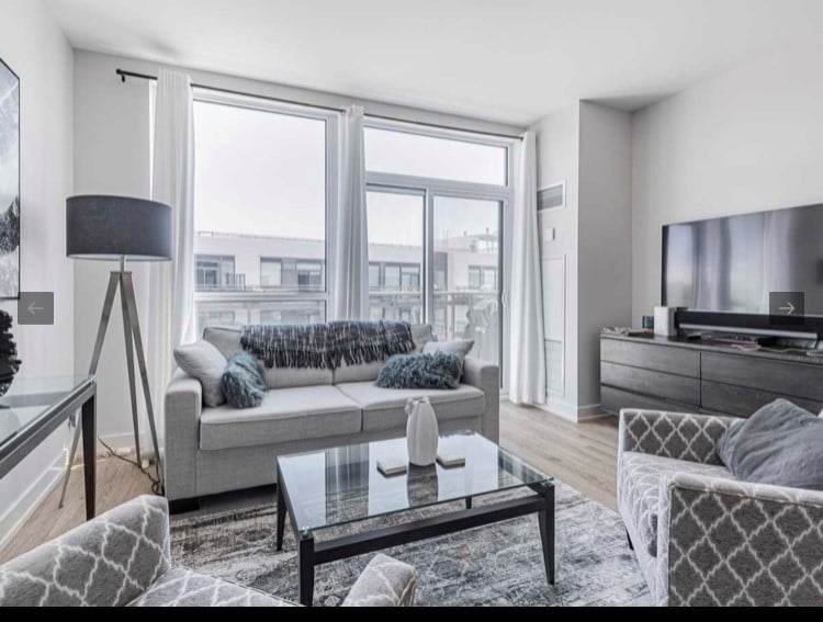 The living area features large windows that allow natural light to fill the space. A light gray sofa is complemented by decorative pillows and a glass coffee table. A flat-screen television is mounted on the wall, while a plush area rug adds texture to the hardwood flooring.