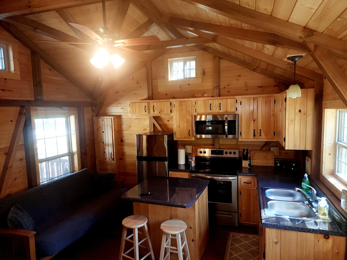 A cozy kitchen area featuring wooden cabinetry and a granite countertop. Stainless steel appliances, including a refrigerator and oven, are visible. A ceiling fan provides comfort while two stools sit near the counter. Light enters through multiple windows, enhancing the warm wood interior.