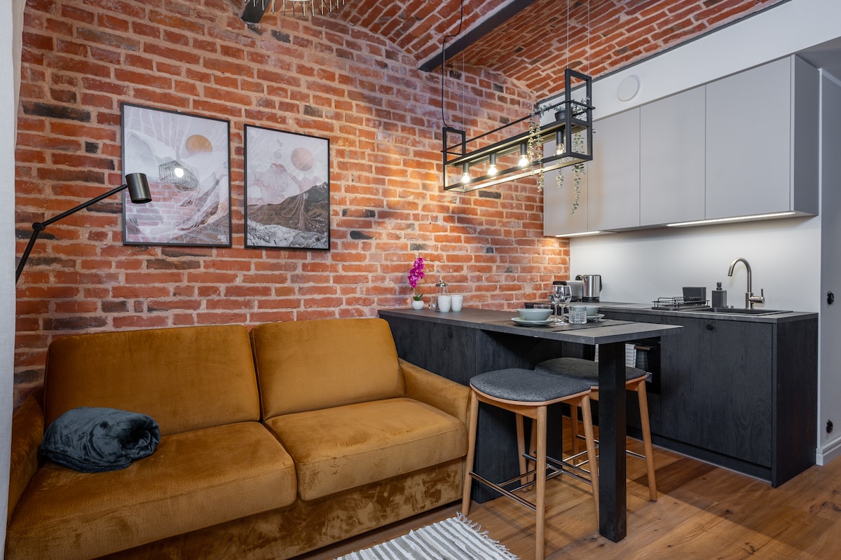 A cozy living space features a rich brown sofa and a dark dining table, accompanied by high stools. Above, a modern light fixture hangs, illuminating the area. The exposed brick wall adds warmth, complemented by framed art pieces and a small flower arrangement on the table.