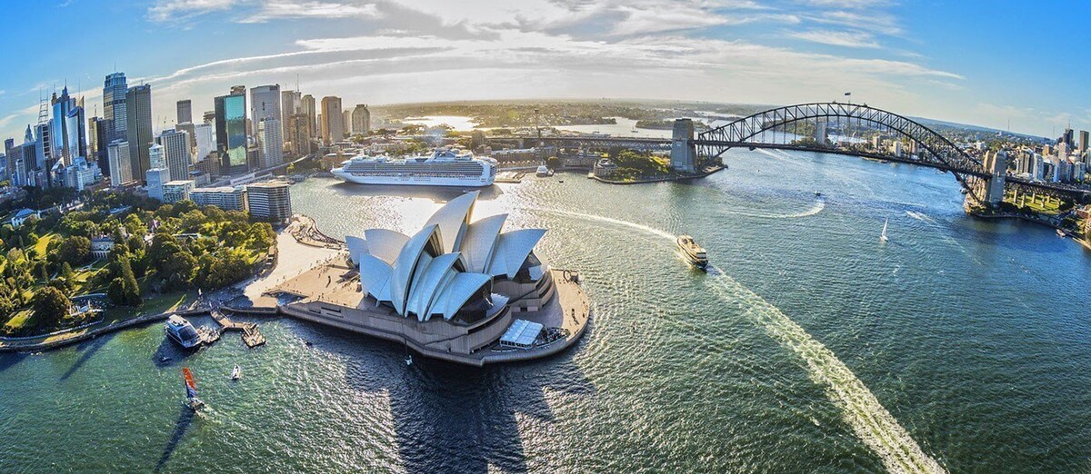 An aerial view captures the iconic Sydney Opera House with its distinctive sail-like design. The image showcases the Sydney Harbour Bridge in the background, with the bustling skyline of the city and boats gently navigating the water, creating a vibrant coastal scene.