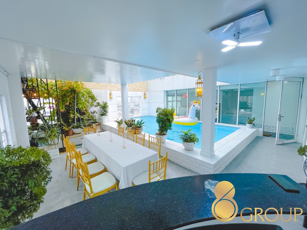 An airy outdoor area features a long dining table surrounded by chairs, with greenery accentuating the space. A bright swimming pool is visible nearby, highlighting a relaxing atmosphere for social gatherings. A ceiling fan circulates air, contributing to the comfortable environment.