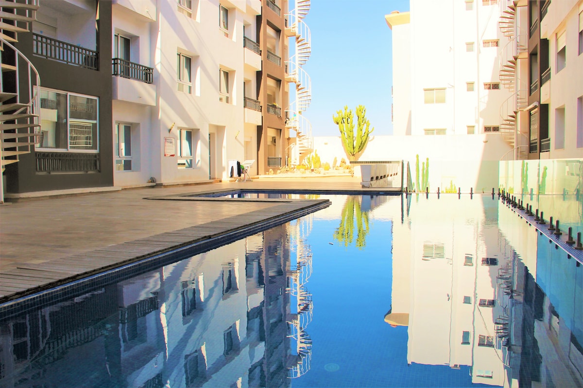 A serene swimming pool reflects the surrounding architecture, bordered by clean, tiled walkways. Sunlight illuminates the area, highlighting the smooth surface of the water. Cacti and vibrant greenery enhance the tranquil atmosphere of the outdoor space.