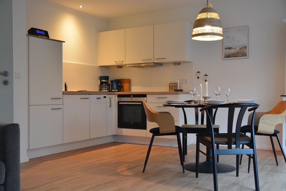 A modern kitchen area features sleek cabinetry and integrated appliances, including a dishwasher and oven. A round dining table with black chairs accommodates four people. Natural light fills the space, highlighting the minimalist design and warm wooden flooring.