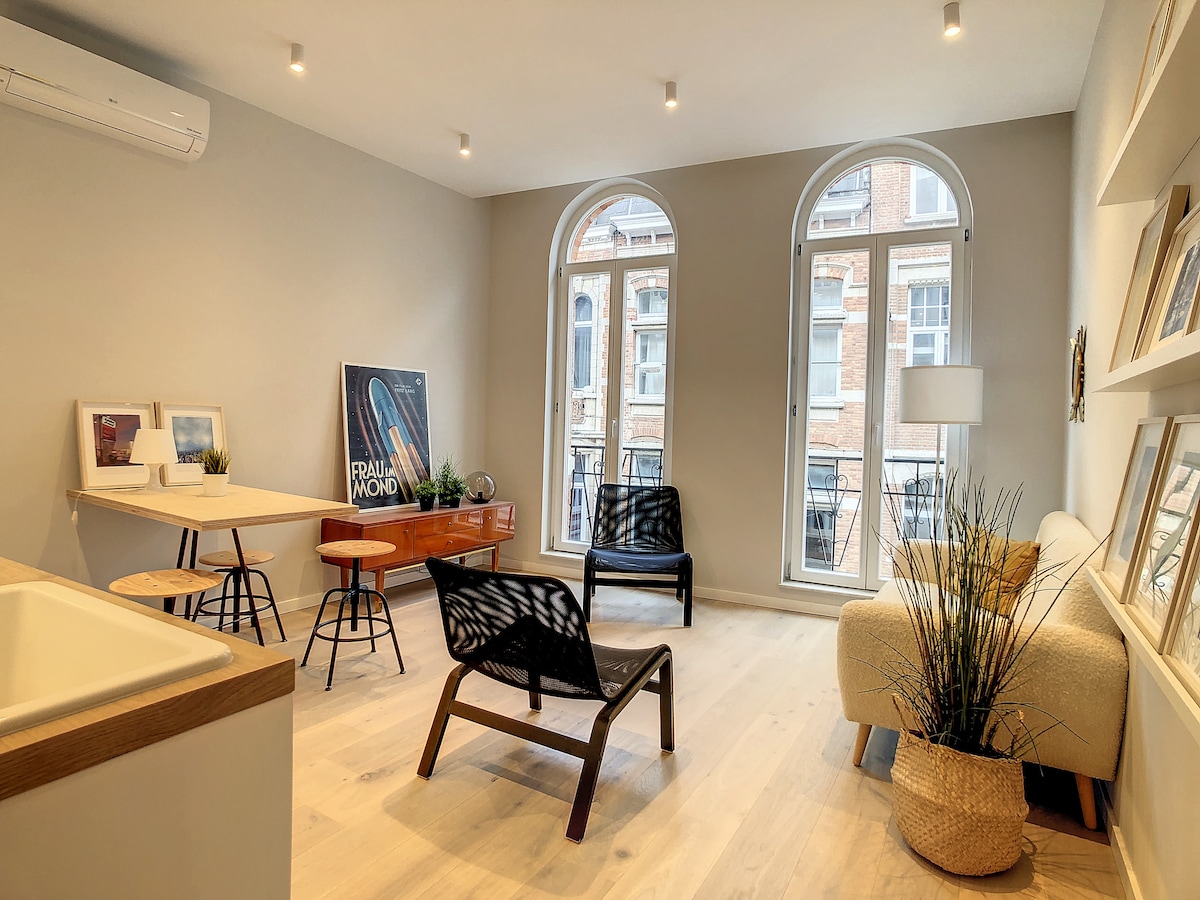 The living area features large arched windows allowing natural light to fill the space. A small dining table with chairs is positioned near the windows, alongside a stylish sideboard. Two black lounge chairs are contrasted by a light-colored sofa, accented by decorative plants.