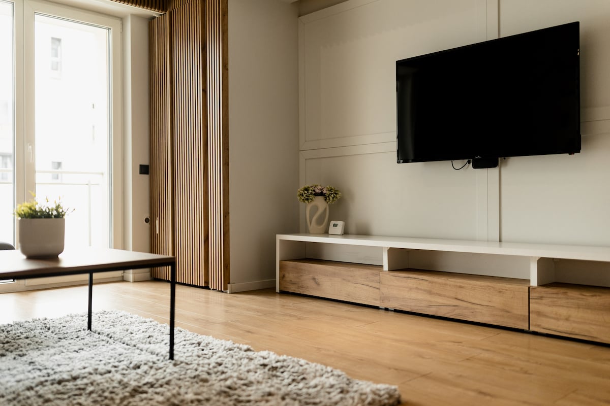 A modern living area features a sleek television mounted against a light wall. A low-profile wooden entertainment unit sits beneath it. A plush area rug covers the floor, and a stylish wooden coffee table is positioned nearby. Natural light enters through a set of glass doors.