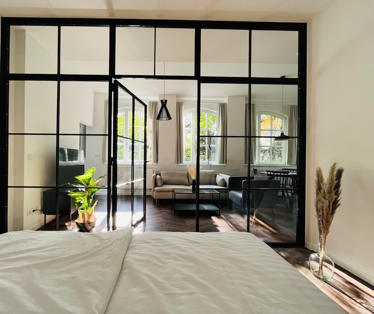 A modern living space is visible through large glass doors. Natural light floods the area, highlighting a comfortable seating arrangement and decorative plants. The bedroom features a cozy bed with white linens, providing a serene connection to the adjoining living area.