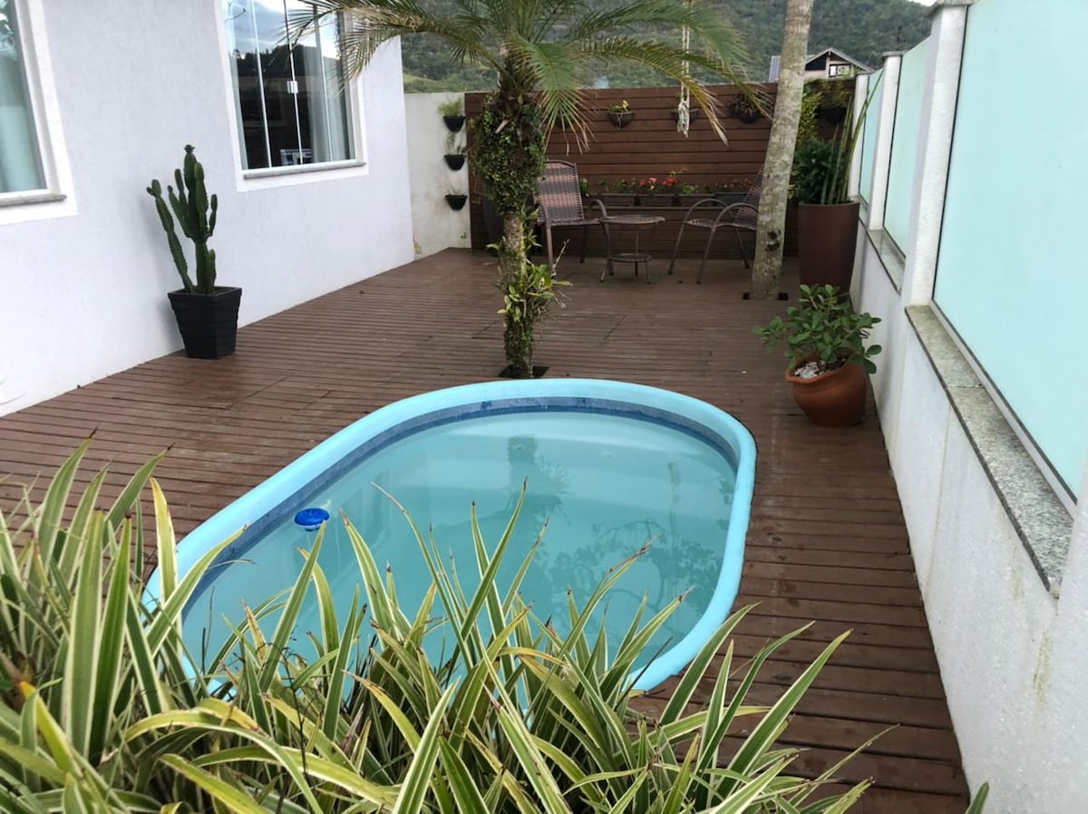 A small pool is surrounded by wooden decking, with potted plants and greenery enhancing the outdoor space. Comfortable seating is arranged nearby, creating a cozy atmosphere. A cactus plant adds an element of interest to one side of the area.