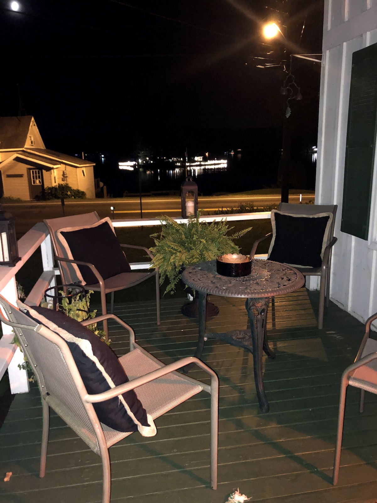 An outdoor porch area is illuminated by soft lighting, featuring four comfortable chairs arranged around a round table. A candle rests at the center of the table. In the background, the calm lake reflects distant lights from across the water.