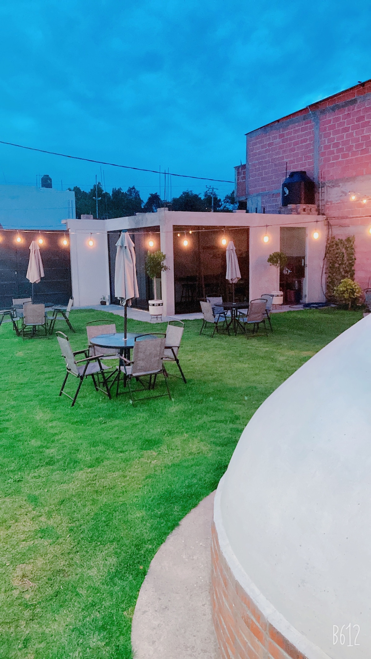 A serene outdoor space features a well-maintained lawn, accompanied by several round tables and chairs. Umbrellas provide shade, while soft lighting enhances the ambiance. A modern structure with large windows is visible in the background, blending with the natural surroundings.