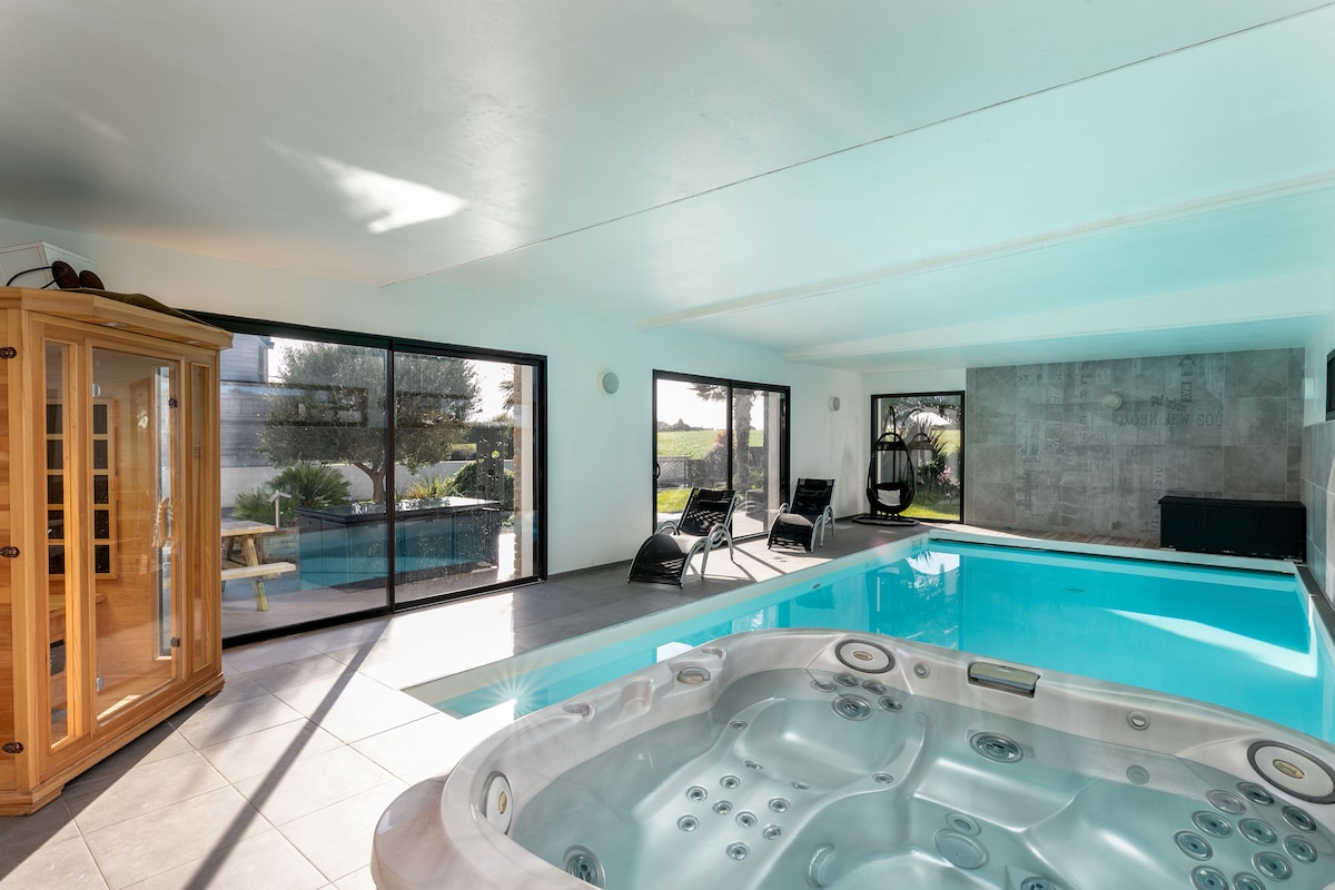 A spacious indoor area features a bright swimming pool and a heated spa. Large glass windows provide garden views and ample natural light, while modern seating creates a comfortable environment for relaxation. A wooden sauna is also visible in the corner of the room.