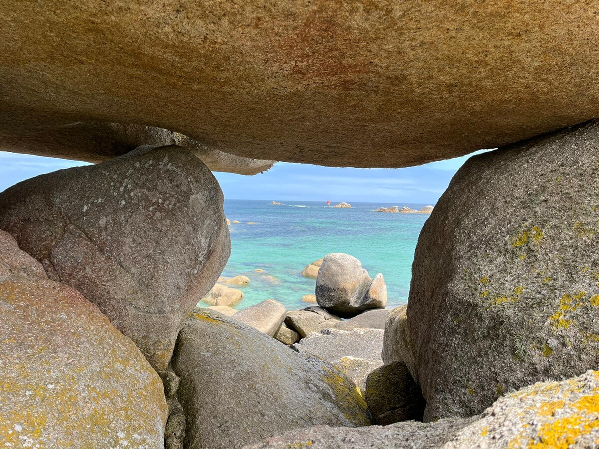 A natural rock formation frames a view of the coastline, with smooth stones and clear turquoise waters visible beyond. The horizon features hints of distant land and soft waves, creating a serene atmosphere of the beach.