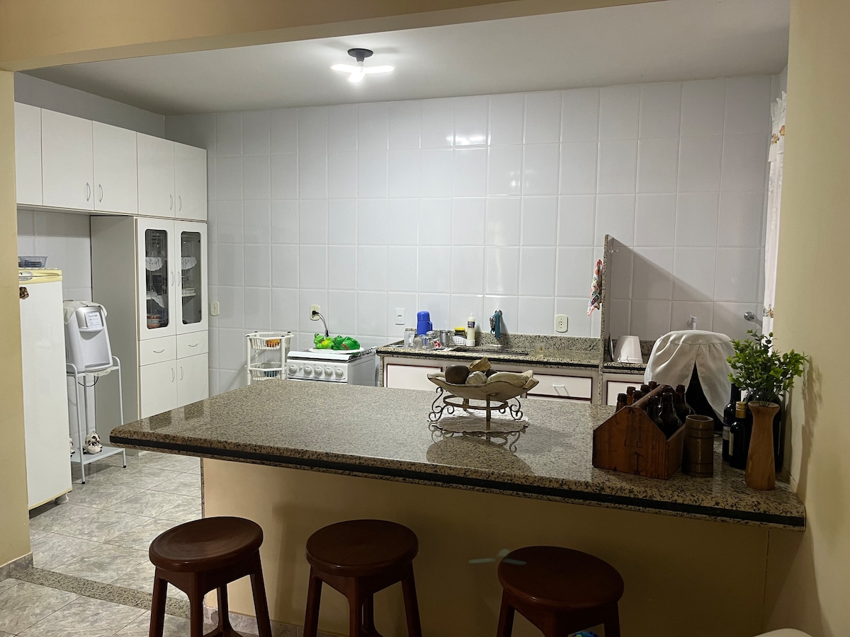 A spacious kitchen features modern cabinetry and a long granite countertop with three wooden stools arranged in front. An assortment of kitchenware is visible, including bowls, utensils, and food items on the counter. Soft lighting illuminates the area, creating a functional cooking space.