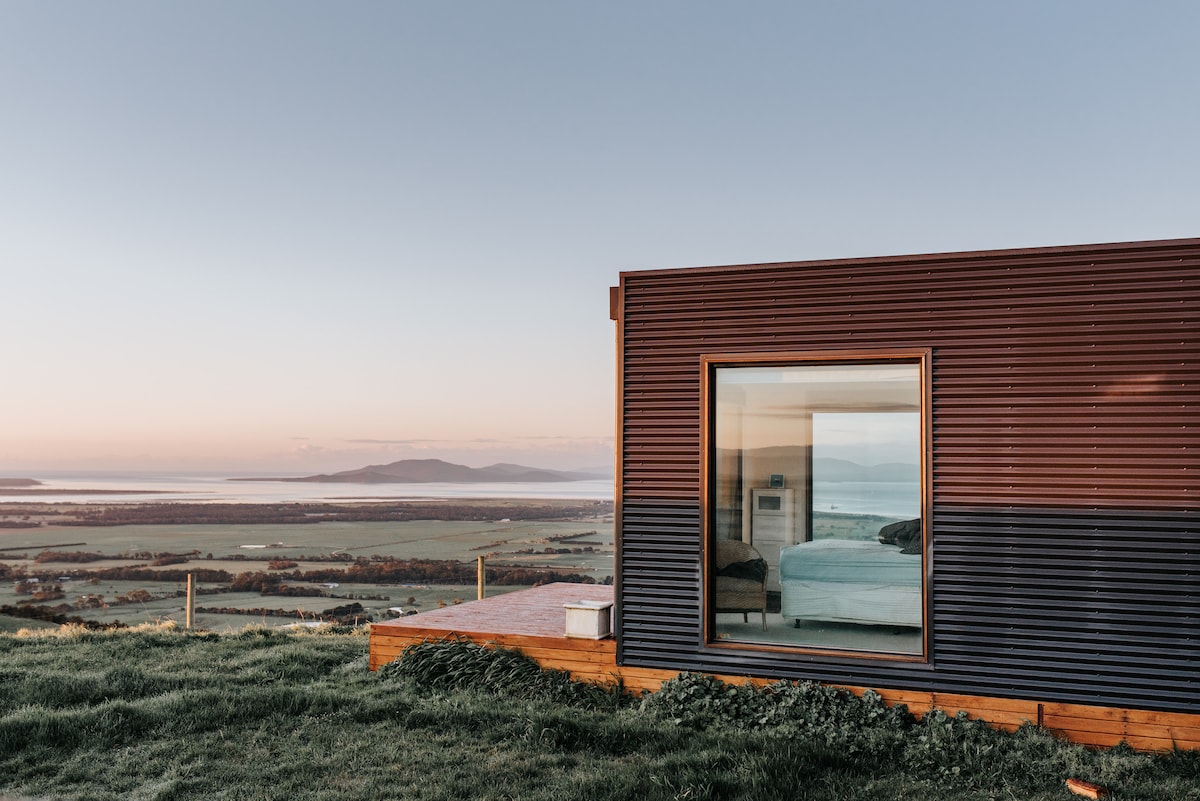 The exterior of a modern villa is highlighted, featuring a large window that frames the serene view of the surrounding landscape. Lush green grass leads to a timber deck that extends from the structure, showcasing the tranquil setting and distant hills.