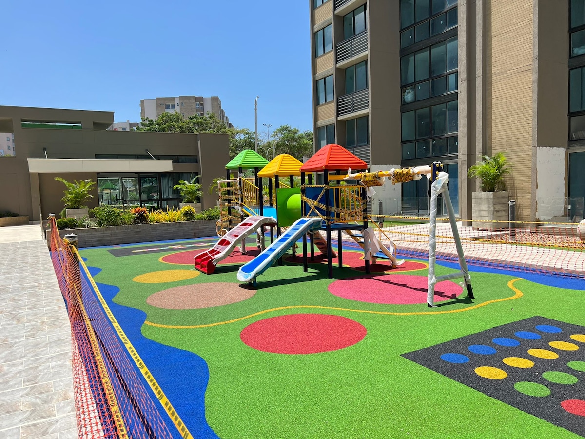 A colorful playground area is highlighted, featuring a play structure with slides and climbing features under shaded roofs. Surrounding the playground, vibrant synthetic turf provides a safe play surface, complemented by playful designs in various colors.