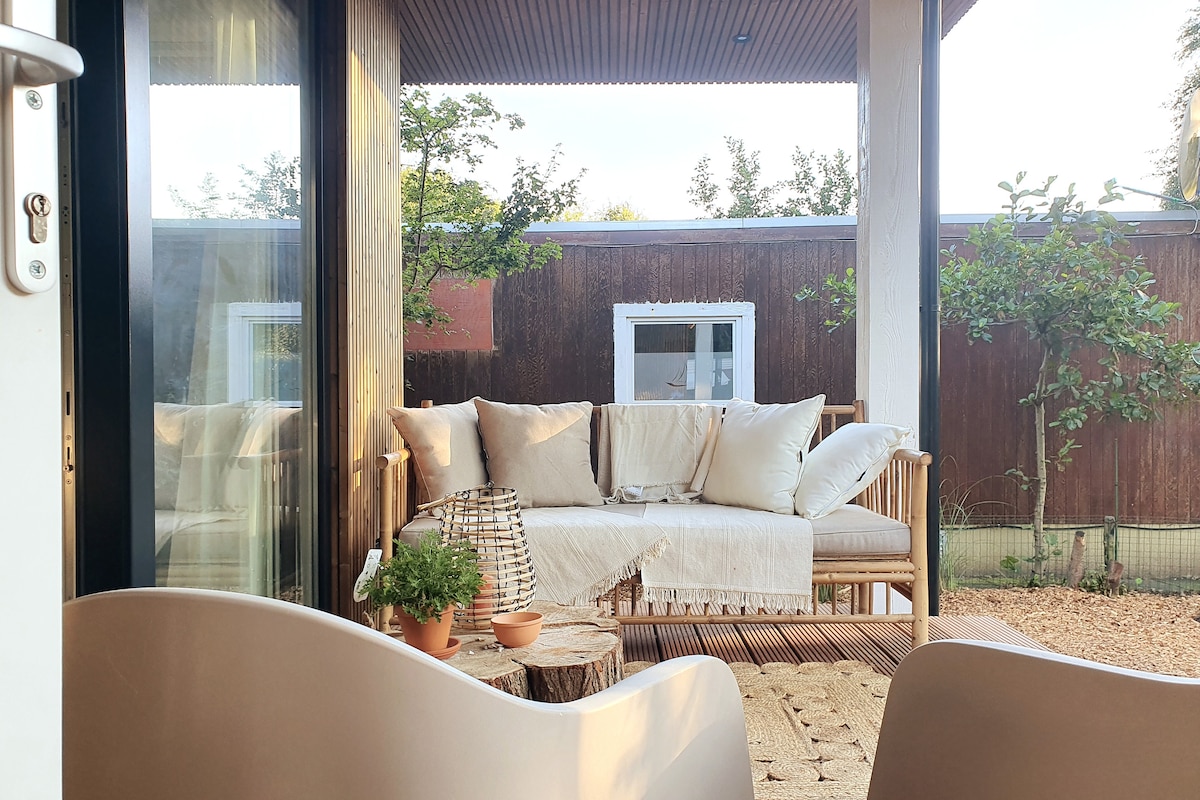 A cozy outdoor seating area features a light-colored sofa adorned with soft cushions. Potted plants are placed on a wooden slatted deck, blending with natural surroundings. Large glass doors allow ample light, creating a serene space for relaxation.