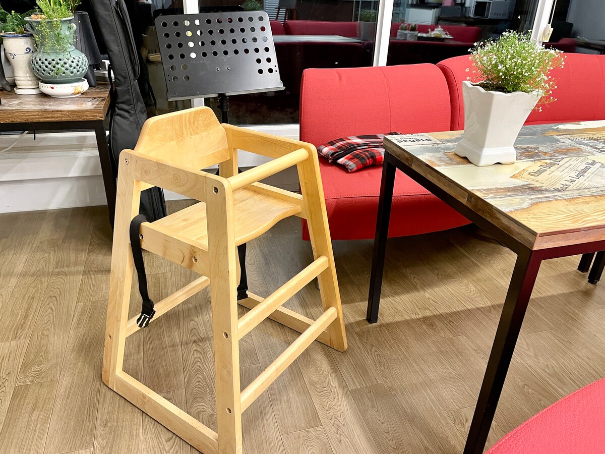 A wooden high chair is positioned next to a rectangular dining table. Red sofas are visible in the background, complemented by a small potted plant on the table. A music stand can be seen nearby, adding character to the inviting interior space.
