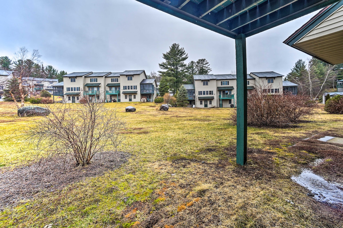 A spacious common area visible from the deck features a lawn with scattered rocks and small shrubs. Townhomes can be seen in the background, framed by trees and cloudy skies.