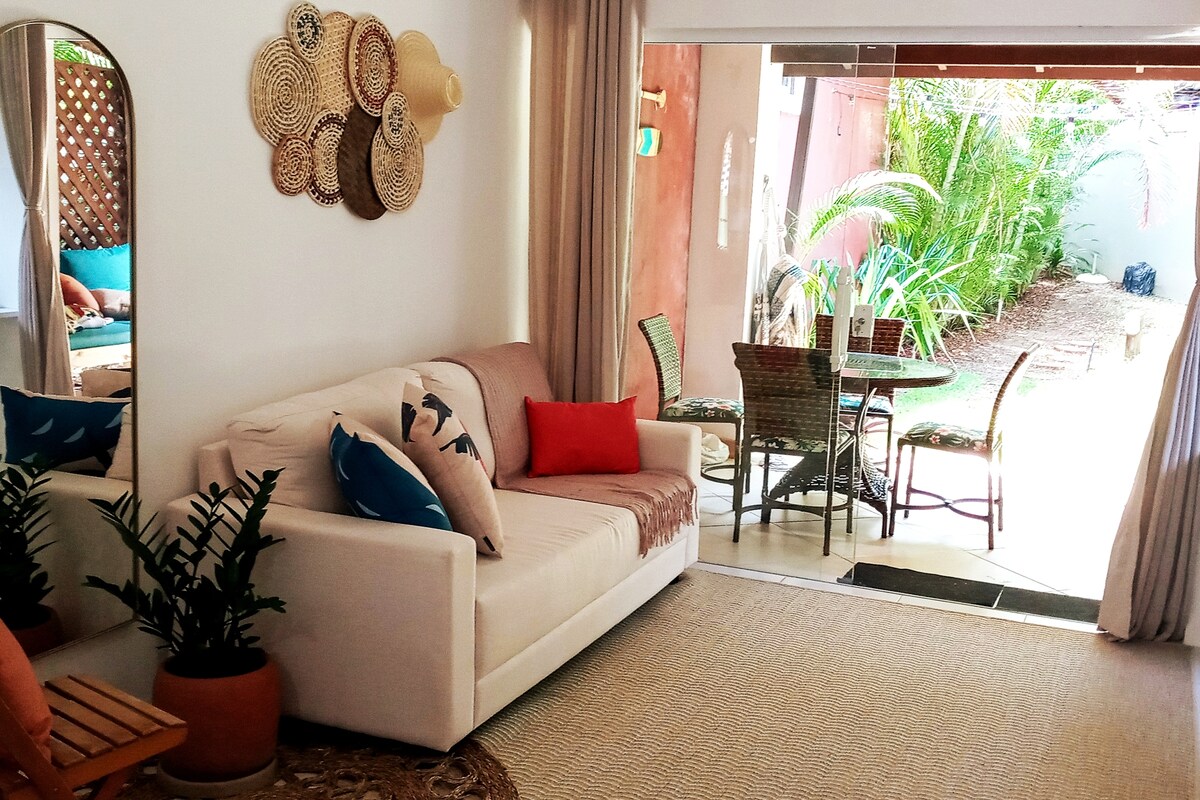 A spacious living area features a comfortable sofa adorned with decorative pillows. Natural light streams through the large glass doors, providing a view of the private outdoor space, which includes a dining table and chairs surrounded by lush greenery.