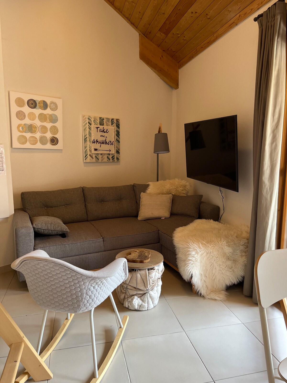 A cozy corner of the living area features a gray sectional sofa adorned with a soft throw pillow and a fluffy rug. A round decorative table sits beside it, and a flat-screen TV is mounted on the wall. Artwork hangs nearby, complemented by natural light from a window.