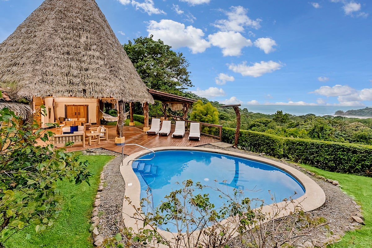 A private pool is surrounded by a spacious deck featuring sun loungers. A thatched roof structure blends into the natural landscape, with lush greenery and scenic views of the surrounding area visible in the background.