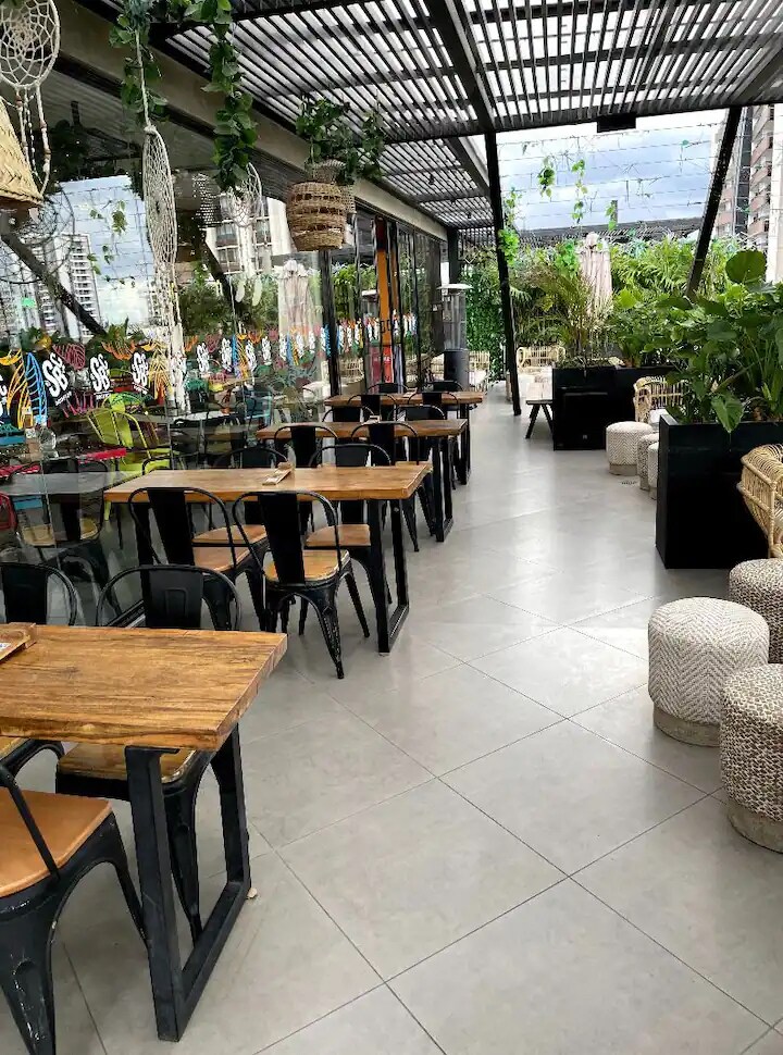 An open terrace dining area is showcased, featuring a row of wooden tables with black metal chairs. Natural greenery surrounds the space, while woven stools add texture. Glass panels provide a view to the outdoors, creating a bright and inviting atmosphere.
