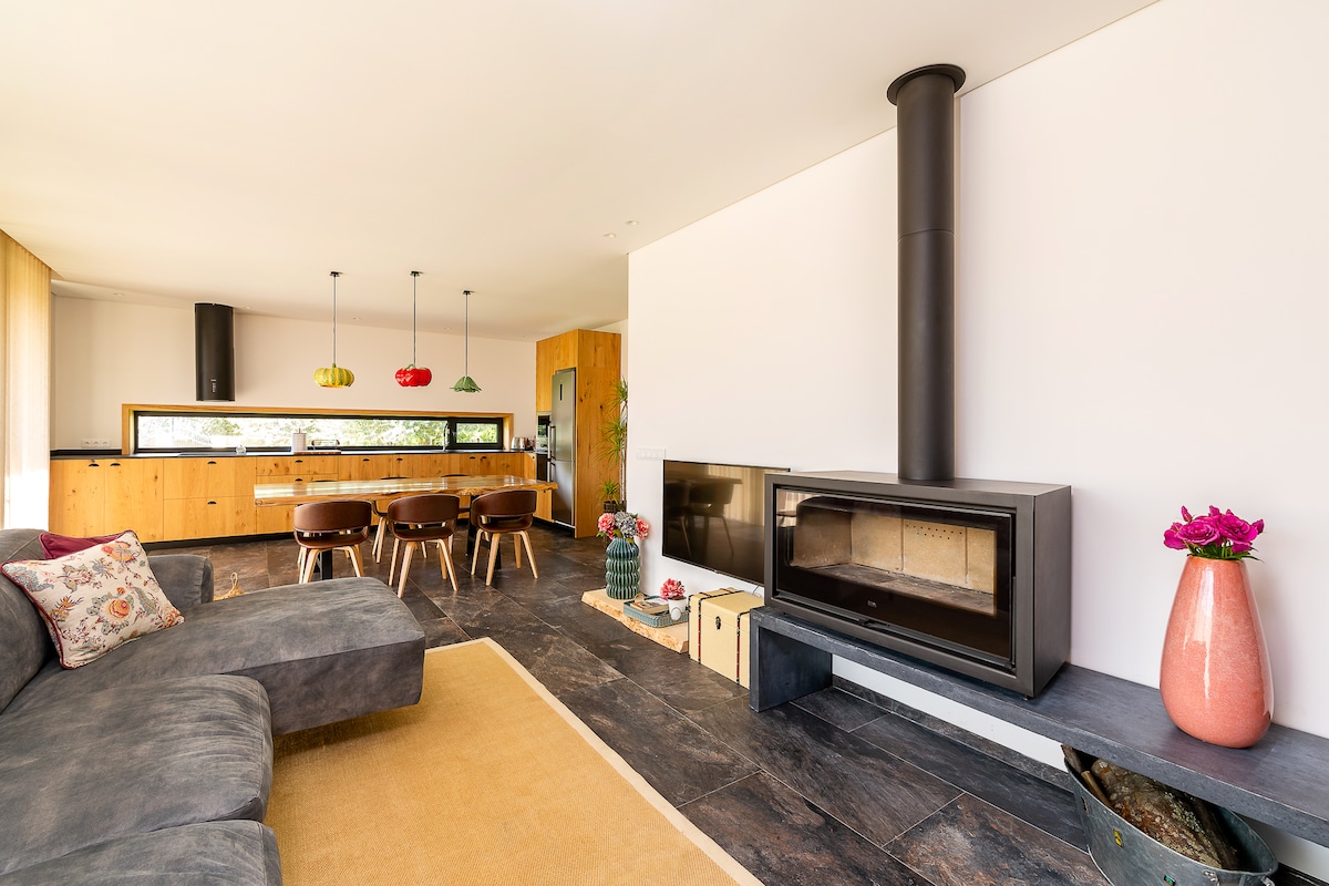 An open living area features a modern fireplace along a sleek stone wall. A comfortable sofa is positioned on a neutral area rug, with bright pendant lights hanging above the adjoining kitchen. Wooden cabinetry complements the overall design, creating a warm and inviting space.