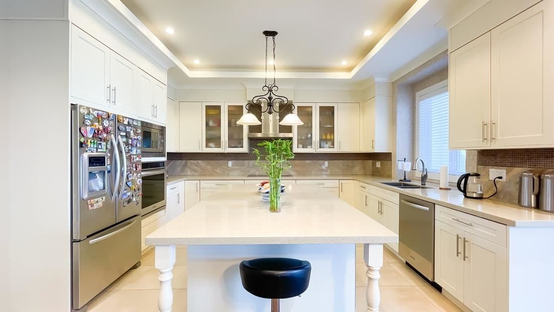 A modern kitchen is displayed with sleek cabinetry and stainless steel appliances. A large central island features a light-colored countertop and is adorned with a vase of fresh greenery. Pendant lights provide illumination above, enhancing the spacious and functional layout.