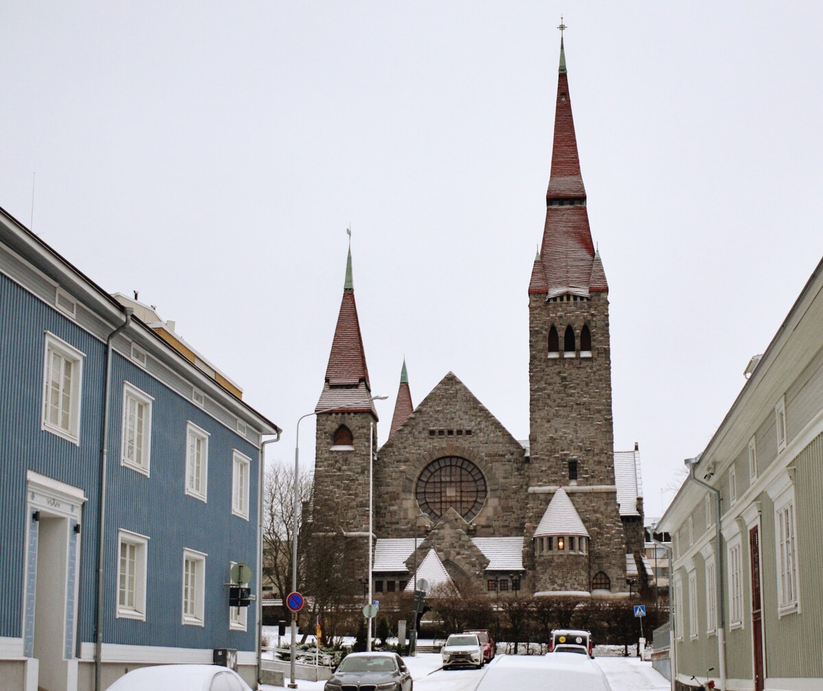 The image captures a snow-covered city scene featuring the prominent stone structure of Tampere Cathedral. The building is characterized by its tall, pointed spires and large, circular stained glass window. Nearby houses in varied colors line the street, creating a charming contrast.