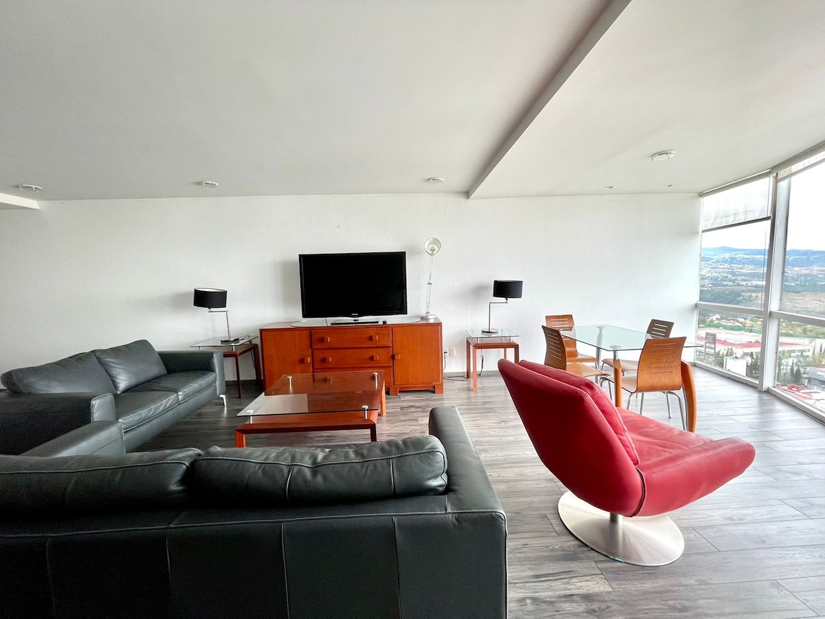 A spacious living area is highlighted by large windows that offer panoramic views. Comfortable seating includes a leather sofa and a vibrant red lounge chair. A wooden side table and a glass dining table are visible, accompanied by dining chairs, creating a multifunctional space.
