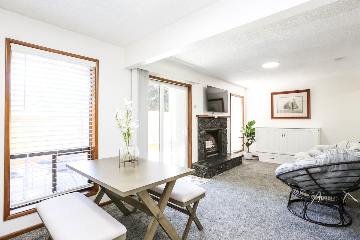 A clean and well-lit studio space features a dining table with benches, a comfortable seating area with soft furnishings, and a fireplace. Natural light floods through large windows, while a small plant adds a touch of greenery. The layout encourages a cozy and relaxing atmosphere.
