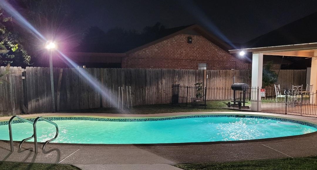A tranquil backyard features a glimmering pool under soft lighting, surrounded by a privacy fence. A patio area is visible, equipped with a BBQ grill and outdoor seating, ideal for relaxing and enjoying meals. Lush greenery can be seen along the edges.