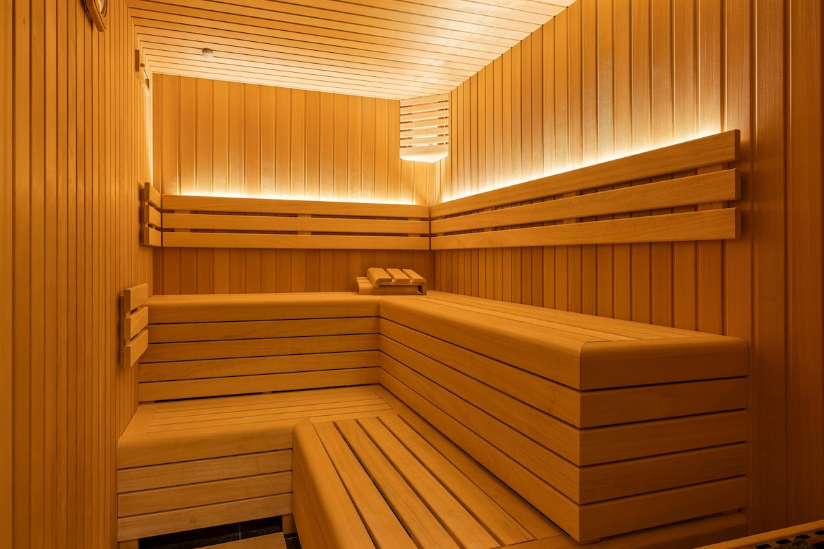 A wooden sauna interior is illuminated by soft lighting along the walls. The space features benches and reclining areas made of smooth timber. The warm tones of the wood create a serene and relaxing environment.