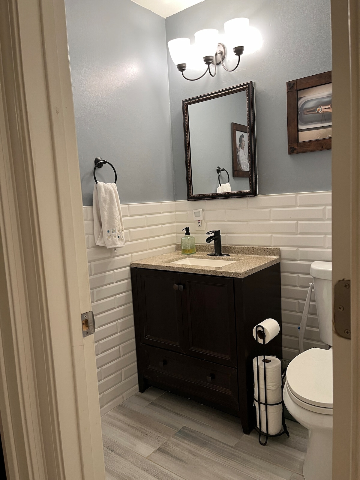 A well-appointed bathroom features a light gray color scheme with white tiled walls. A dark wooden vanity with a granite countertop holds a sink and soap dispenser. A mirror is mounted above the vanity, and a framed picture adds visual interest. Essential amenities, including towels and toilet paper, are neatly arranged.