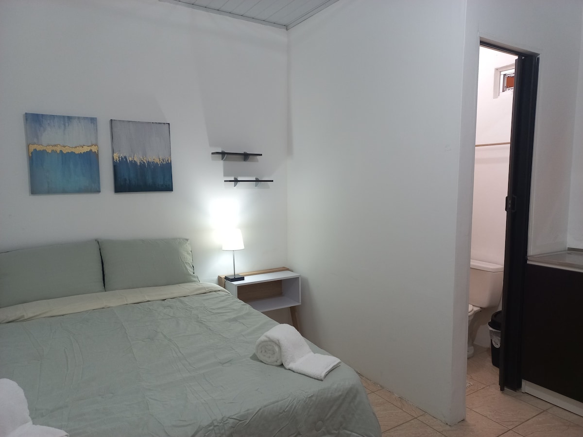 A spacious room features a comfortable bed with light green bedding. Two abstract paintings with blue and gold hues adorn the wall above the bed. A small nightstand with a lamp provides soft lighting, while a doorway leads to a compact bathroom within the room.
