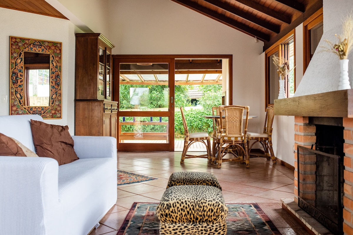 A bright living area features a white couch adorned with brown pillows and a patterned ottoman. Large windows provide views of the lush outdoor space, while a brick fireplace adds a cozy focal point. Wooden accents and a decorative mirror complement the home's inviting atmosphere.