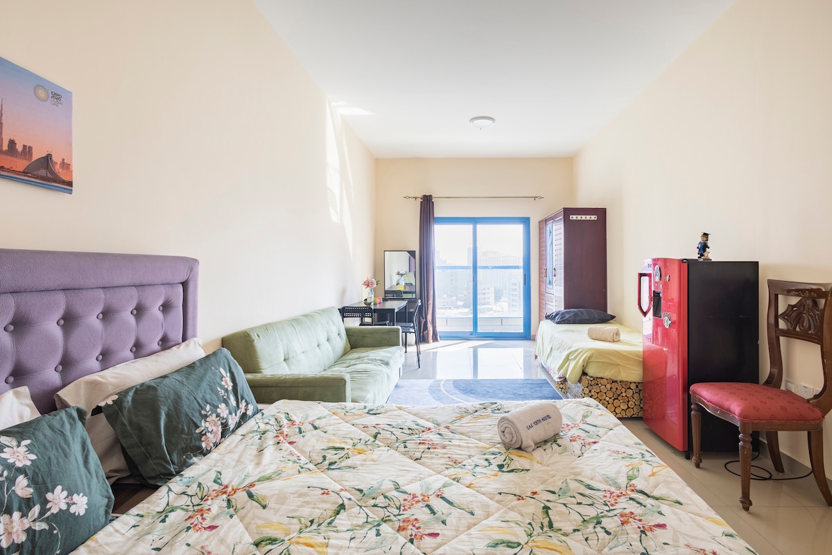 A spacious room is equipped with multiple sleeping areas, featuring a vibrant quilt on one bed and decorative pillows. A green sofa provides additional seating, while a glass door offers views of the outside. A red refrigerator and a small table are also present in the room.