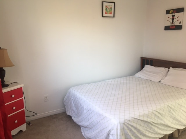 A comfortable double bed is placed against a light-colored wall, with neatly arranged white linens. A nightstand with a lamp and a small red dresser add color to the room. Light from the window highlights the simple and functional design.