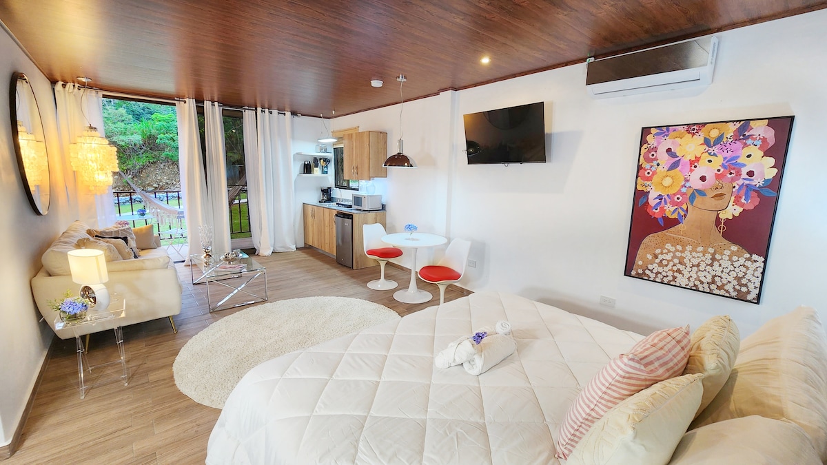 A bright and modern studio suite showcases a spacious bed with a quilted cover and decorative pillows. A cozy seating area is complemented by a glass topped table with red stools. The kitchenette area features wooden cabinetry. A smart TV is mounted on the wall, and light fixtures add warmth.