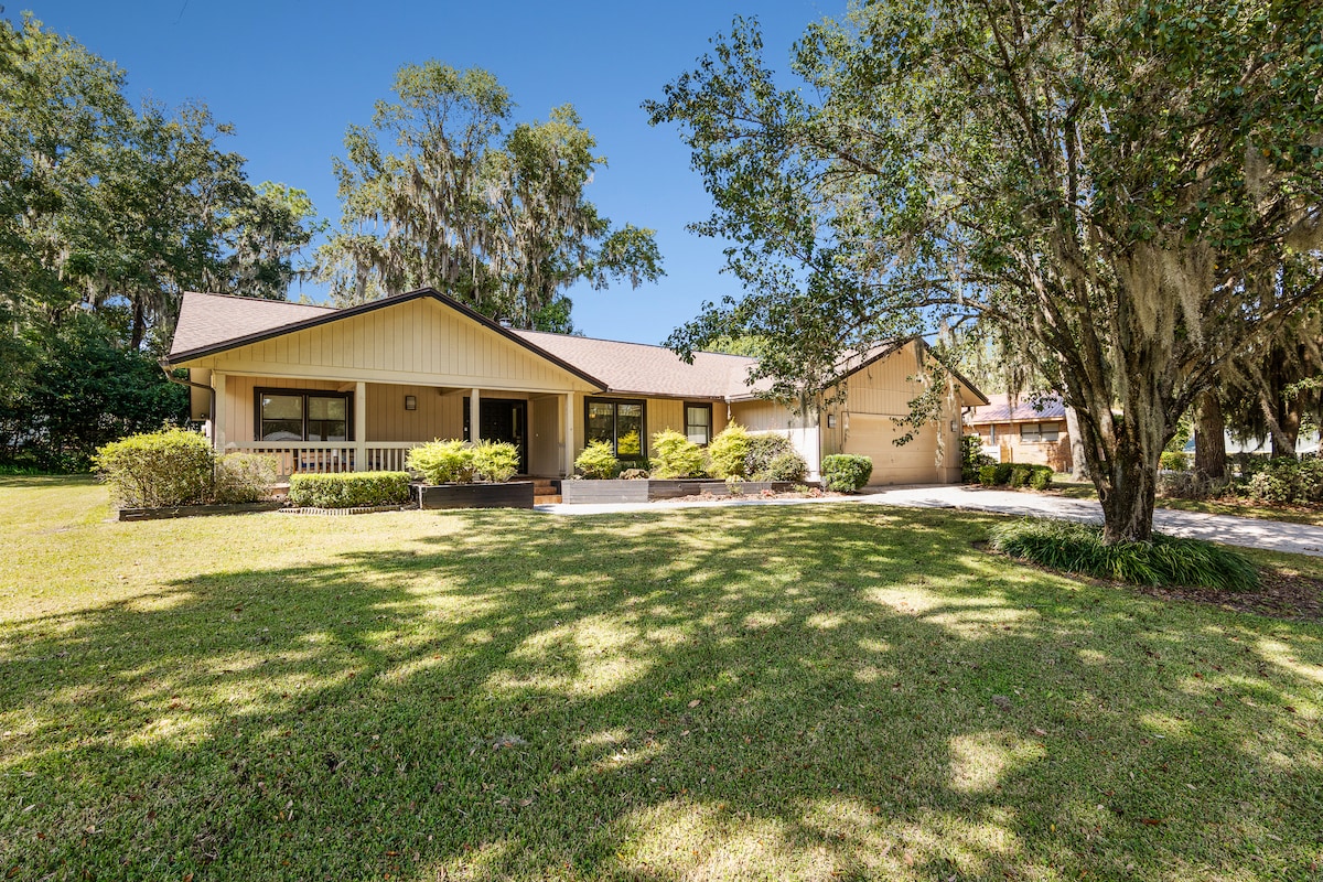 A single-story home is surrounded by a green lawn and mature trees. The facade features a covered porch with seating and a light-colored exterior. A garage is visible on the right side of the property, contributing to the overall peaceful ambiance of the location.