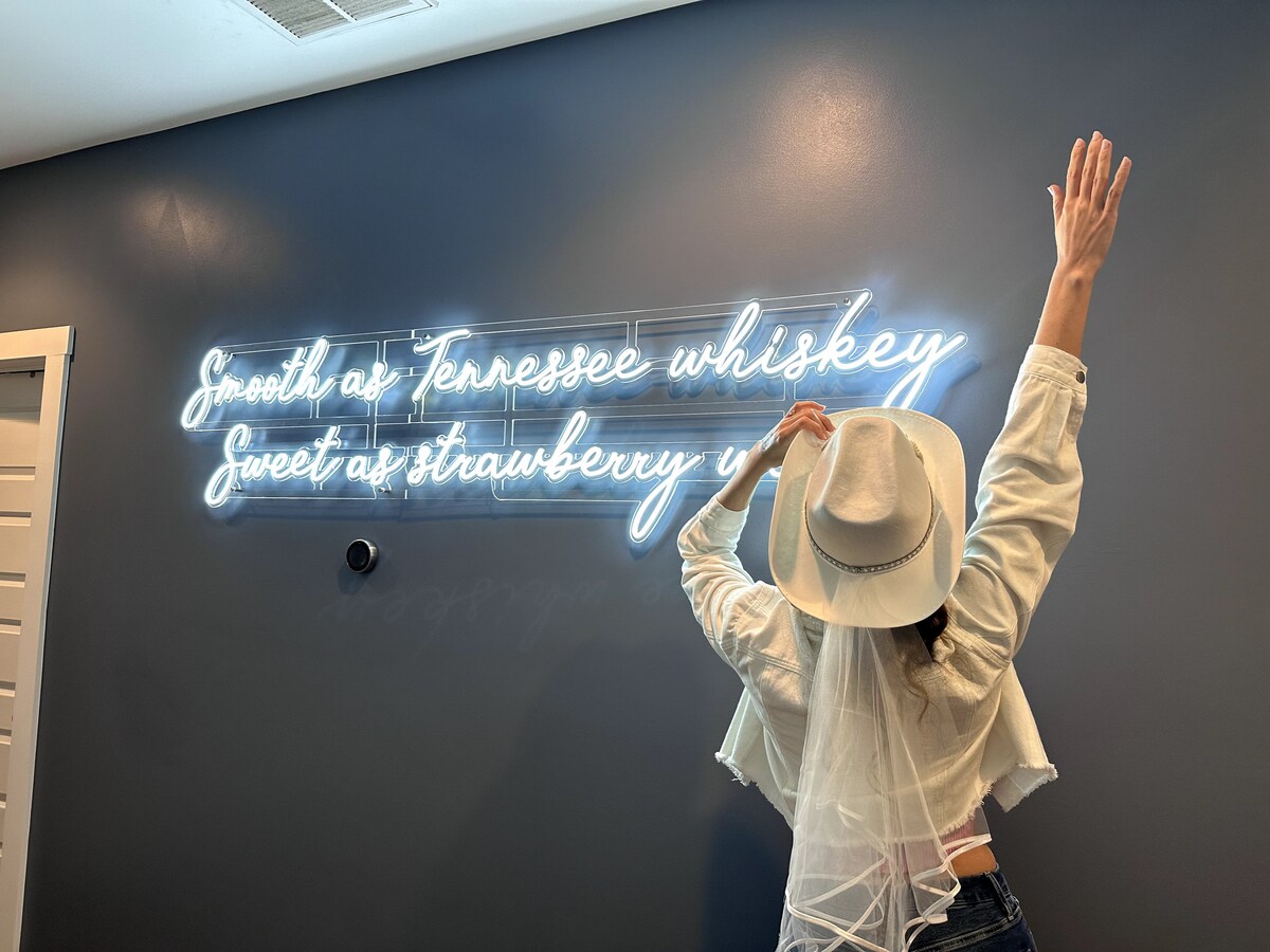 A neon sign featuring the phrase 'Smooth as Tennessee whiskey, Sweet as strawberry wine' is displayed on a dark blue wall. A person wearing a white hat and veil poses beneath the sign, creating an engaging Instagram photo opportunity.