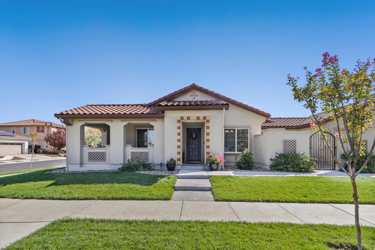 A welcoming facade of a single-story home features a tiled roof and decorative elements. The front path is lined with green grass and flowers, leading to a dark front door framed by large windows on either side. A well-maintained landscape enhances the entrance.