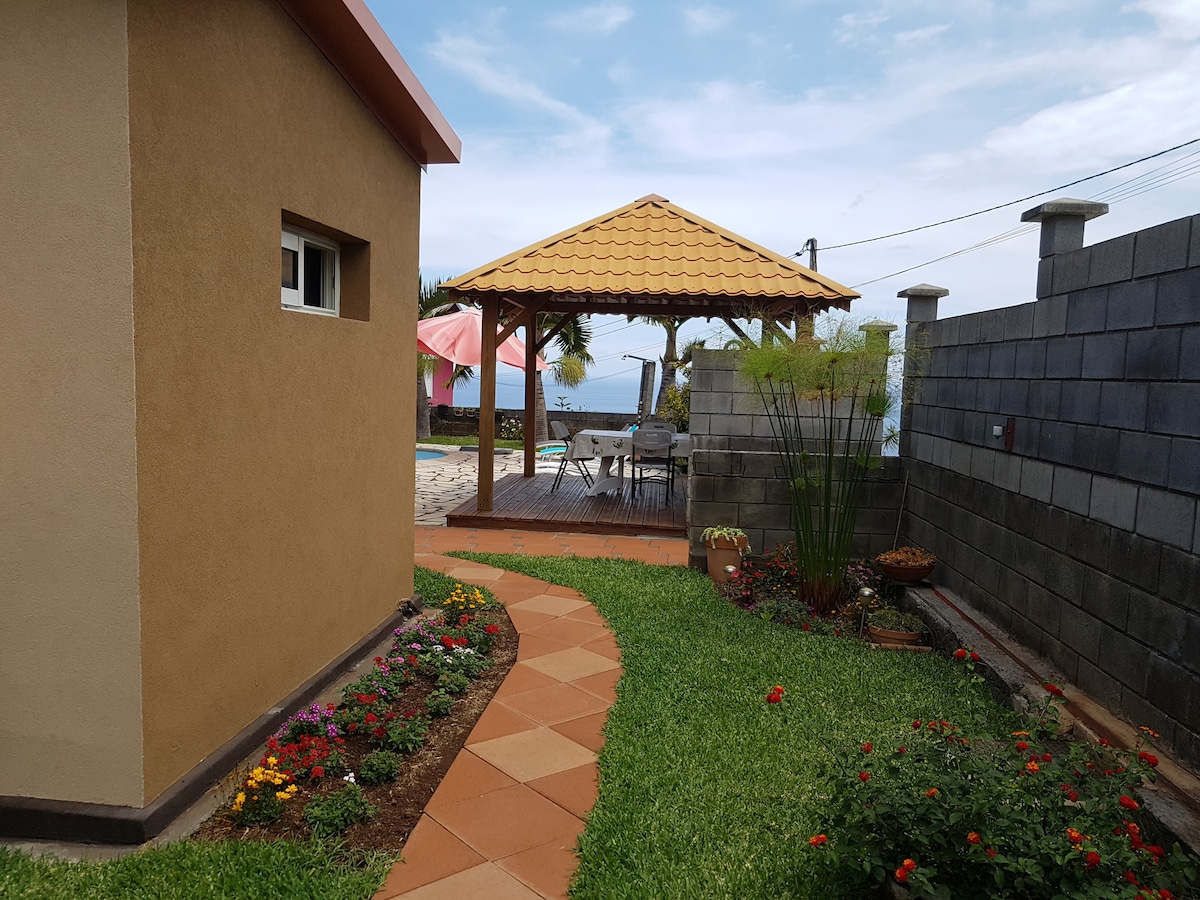 A pathway lined with colorful flowers leads to a wooden gazebo with a tiled roof, situated near a swimming pool. The surrounding greenery provides a serene atmosphere, with a small building visible to the left and a stone wall framing the area.