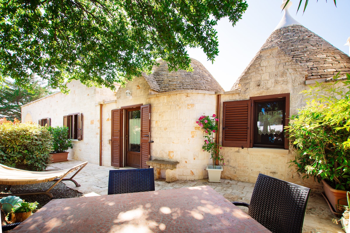 The exterior of the trulli accommodation is shown, featuring traditional stone architecture. Lush greenery surrounds the area, and a patio with a table and chairs is visible. Large windows with wooden shutters add charm, while a hammock and potted plants create a relaxing outdoor space.