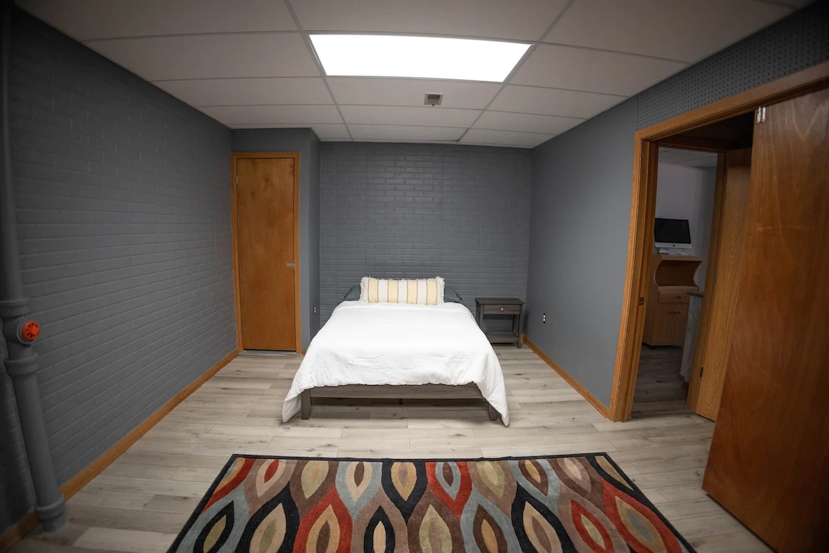 A spacious sleeping area features a large bed with a white duvet and colorful accent pillows. Light wood flooring is complemented by a patterned area rug. Neutral gray walls provide a calm ambiance, while a wooden door and an open doorway lead to additional spaces.