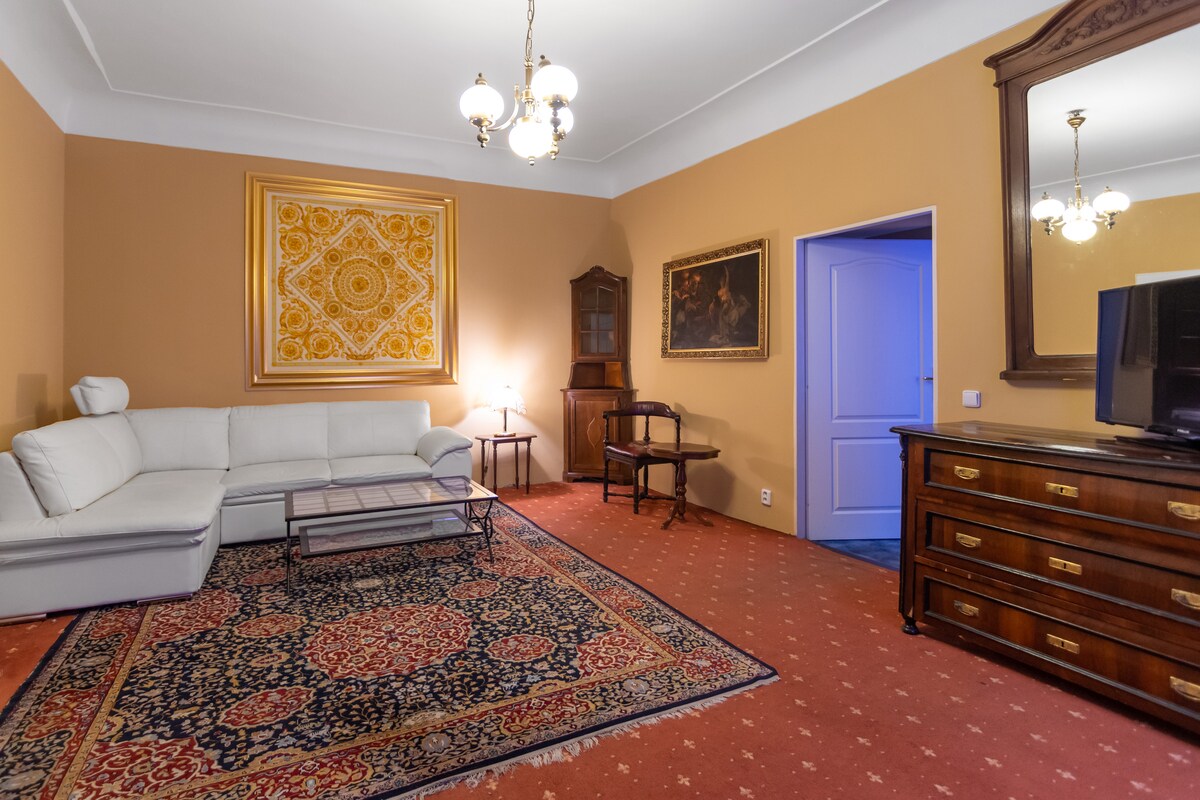 A spacious living area features a large white sectional couch and a glass coffee table. An ornate area rug with intricate patterns covers the floor. A tall cabinet and a vintage-style chair are present, illuminated by warm lighting from ceiling chandeliers.