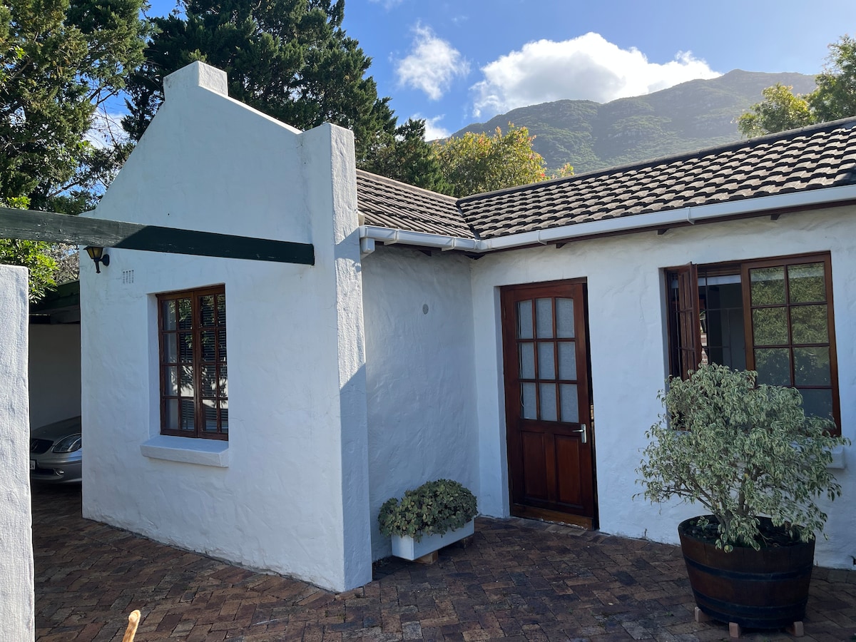 A charming exterior features whitewashed walls with a traditional roof. Two wooden doors are highlighted, surrounded by windows on either side. A small potted plant adds greenery to the entrance, while a paved area provides space for parking.