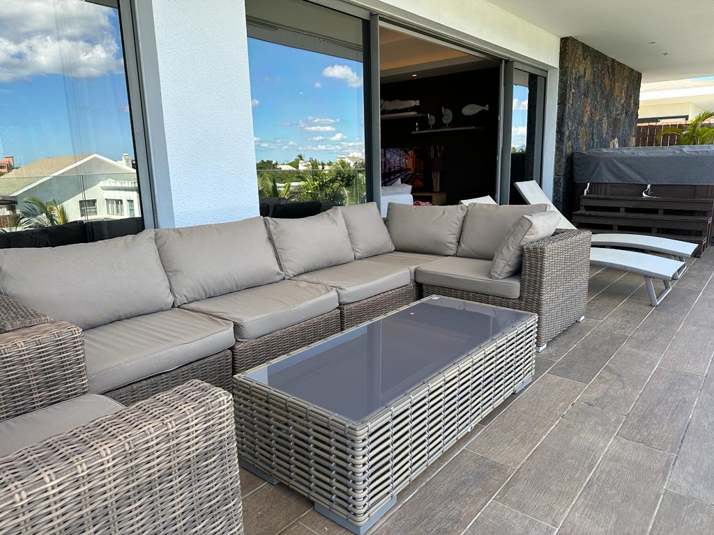 A spacious outdoor seating area features a large sectional sofa with beige cushions, complemented by a glass-top coffee table. Large sliding glass doors provide access to a bright interior space, while the surrounding tiles contribute to a modern and inviting atmosphere.