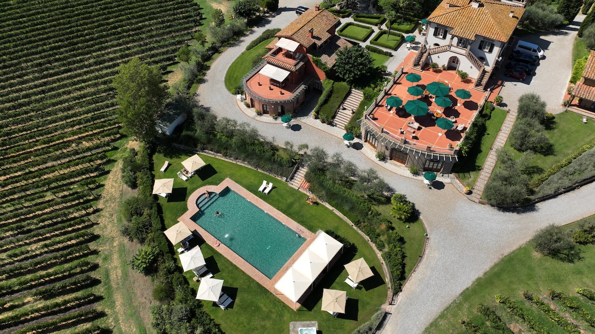 An aerial view showcases a spacious outdoor area featuring a rectangular pool surrounded by lounge chairs and shaded cabanas. The property includes various structures, with terracotta roofs and green umbrellas on a terrace. Lush vineyards extend into the background, enhancing the serene landscape.