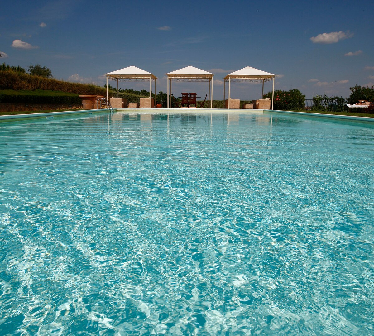 A large swimming pool reflects the sky, surrounded by well-maintained green spaces. Two shaded cabanas are positioned along the pool's edge, offering a spot for relaxation. Lounge chairs are visible nearby, inviting guests to unwind in the serene outdoor environment.