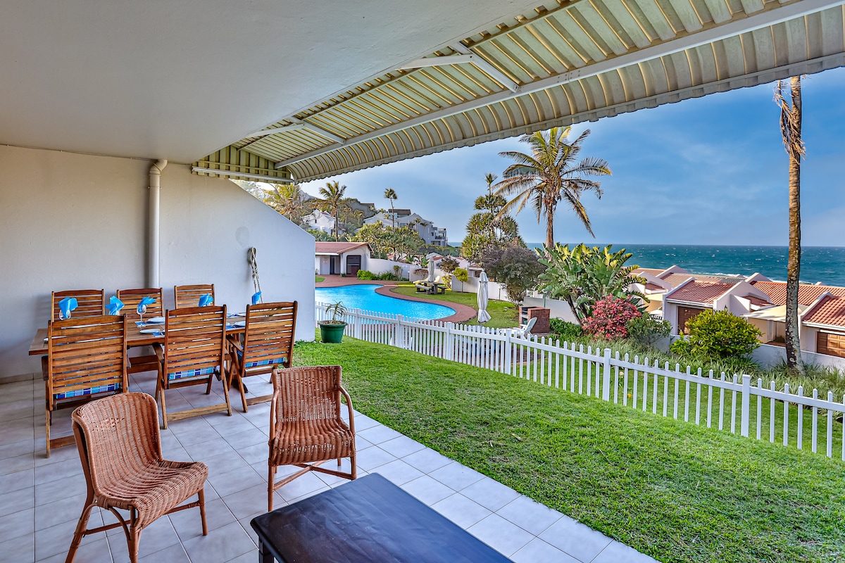 An outdoor terrace features a dining table with wooden chairs and a coffee table. Lush green grass surrounds the area, with a view of the swimming pool and the ocean beyond. Palm trees are visible, indicating a tropical atmosphere and creating a relaxing setting.